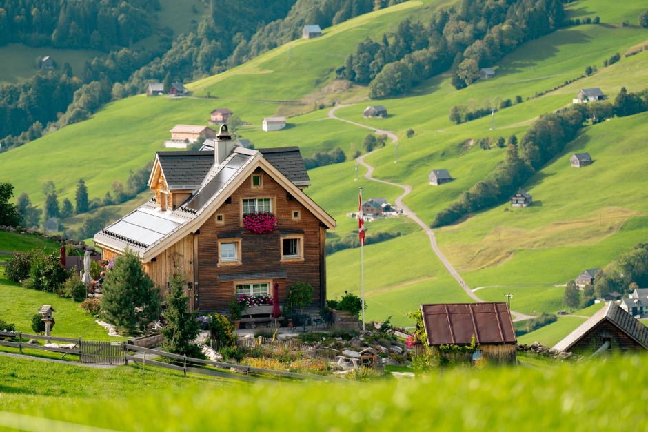 A charming wooden house with a grey tiled roof is situated in a lush, green hillside landscape. The house features a small front garden with vibrant flowers, shrubs, and a decorative hanging basket of bright pink blooms beneath the window. To the right of the house, there is a weathered metal shed with a rusted brown roof, positioned on a grassy slope. A flagpole displaying a red and white Union Jack flag stands nearby, indicating a possible UK location. Surrounding the property, the rolling terrain is covered in well-maintained grass and scattered trees, with a low wooden fence bordering the garden area. In the background, multiple small houses are scattered across the hillside, connected by winding gravel paths that traverse the vibrant green landscape. The scene is bathed in natural daylight, highlighting the vivid colors and textures of the rural setting, with no visible waste or rubble in the vicinity, emphasizing the peaceful and tidy environment typical of private rural homes near countryside rubbish collection services or alternative disposal options.