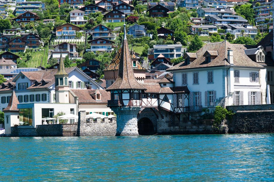 A scenic lakeside view featuring a historic European-style building with a pointed, tiled roof and a small turret, situated directly along the water's edge. The building has white plastered walls with dark wooden beams, and several windows with lattice-style shutters. Adjacent to the building, a stone and brick structure extends into the water, incorporating a small arched passageway beneath the turret. In the background, there are numerous modern residential houses of varying sizes and styles positioned on a hillside, surrounded by lush green trees and vegetation. The water in the foreground reflects the bright, clear sky, and the overall scene presents a peaceful, picturesque environment. This setting illustrates a location where private or alternative waste management services might be relevant, with the emphasis on the integrity of the historic architecture and natural surroundings.