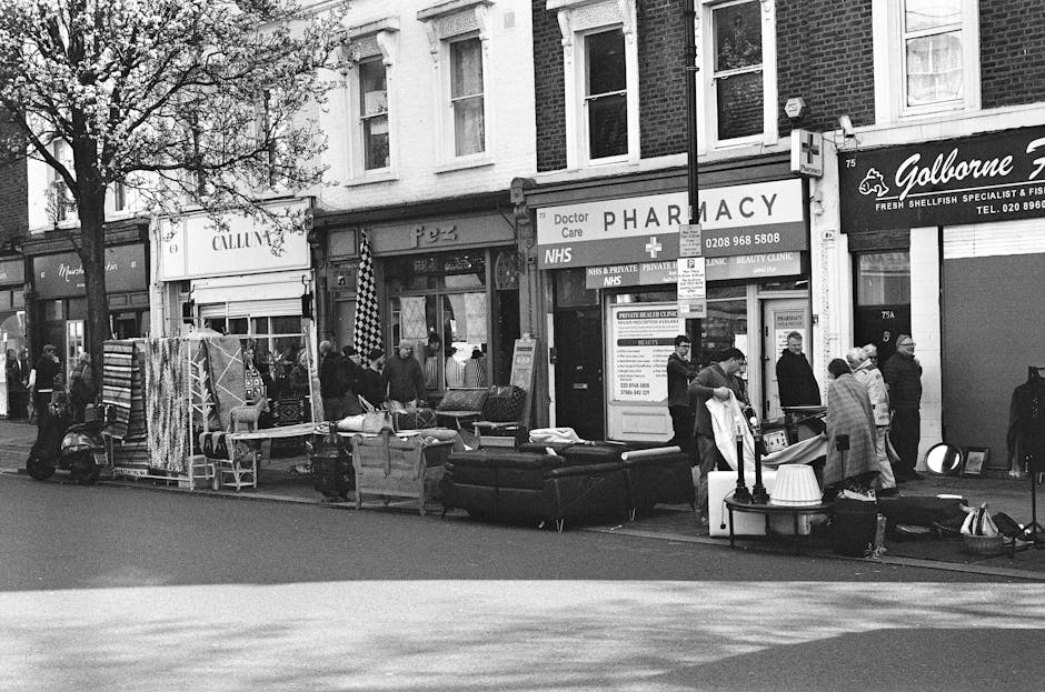 A street scene outside a row of shops and a pharmacy, with several groups of people gathered around an assortment of discarded furniture and household items. Visible objects include a dark upholstered sofa, a few wooden chairs, a small table, and several boxes or containers, all arranged along the pavement in front of the storefronts. The furniture appears to be used and slightly worn, with textures ranging from the smooth fabric of the sofa to the rougher wooden surfaces of the chairs. The items are situated close together, some leaning against each other, indicating they are intended for collection or disposal. In the background, shop windows with signage, including a pharmacy with NHS and private care notices, are partially obscured by a streetlamp and a parking restriction sign. A scooter is parked nearby, and pedestrians are walking or standing on the sidewalk, some engaged in conversation or examining the items. The overall scene is captured in natural daylight, with shadows cast across the pavement, illustrating a typical urban environment where private waste clearance or alternative rubbish removal services might be utilized, such as those provided by wastecollectionswisscottage.co.uk, to handle bulky waste on-site outside retail or residential premises.