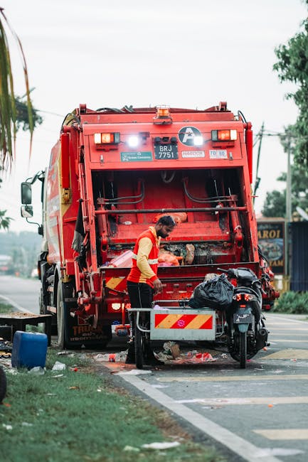 A waste collection operative wearing a high-visibility yellow and red jacket is seen standing at the rear of a large, red garbage truck on a paved urban street. The truck is equipped with hydraulic lifting mechanisms and has a white and orange reflective chevron pattern on its rear section. The worker appears to be using tools or inspecting the load, which includes a black plastic bag and other waste materials. The surrounding environment features a sidewalk with a patch of grass, a blue container, and some scattered debris. In the background, there are blurred trees, power lines, and buildings, indicating a semi-urban setting. The scene is illuminated by natural daylight, suggesting a typical day for private waste collection or rubbish removal services, consistent with alternative waste handling methods such as on-site clearance or independent collection by [COMPANY_NAME]. The overall composition emphasizes the practical aspect of waste disposal, with focus on the vehicle, materials, and worker activity.