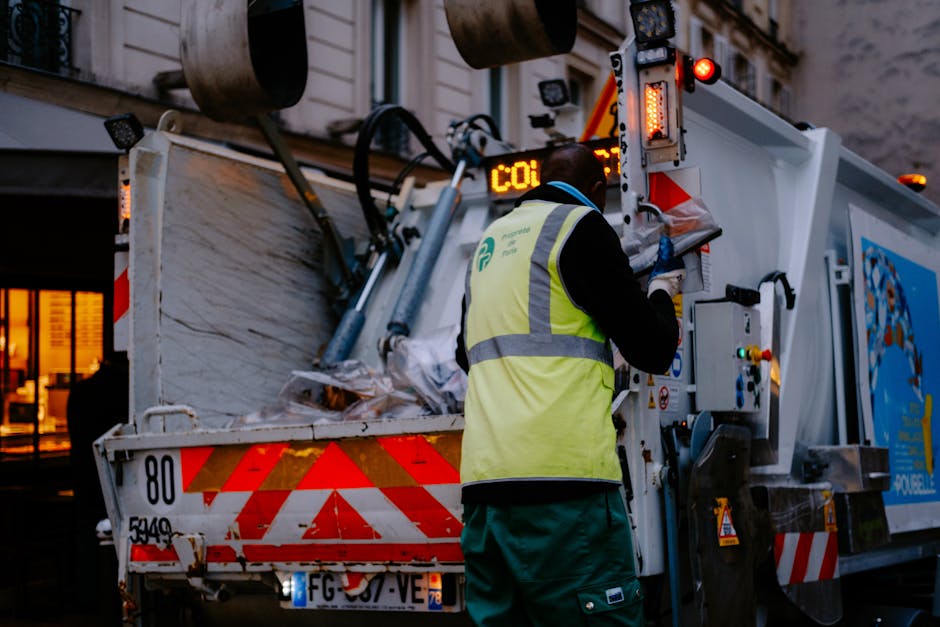 A waste collection worker, wearing a high-visibility yellow vest with reflective stripes and blue gloves, stands at the rear compartment of a commercial rubbish collection vehicle during dusk. The vehicle is white with red and orange reflective markings along its sides and rear, and features bright lights illuminating the work area. Inside the open container, various scrap materials are visible, including crumpled paper, plastic, and other debris. The worker appears to be operating controls or recording information on a digital display panel attached to the vehicle. In the background, a building with lit windows suggests an urban environment, and the scene emphasizes efficient waste management or private rubbish removal services in an outdoor setting, potentially relating to on-site clearance or alternative disposal methods.