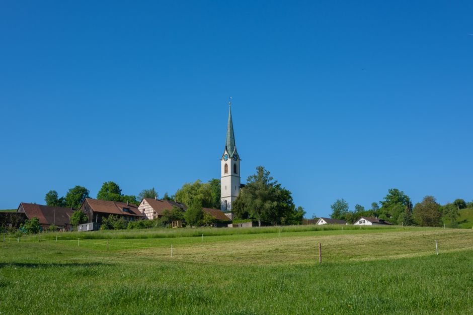 A rural landscape featuring a small village with several houses and buildings situated on a gentle grassy hillside. The houses have sloped roofs, some with visible red or brown tiles, and are surrounded by green trees and bushes. In the background, a prominent white church with a tall, slender, pointed steeple topped by a cross rises above the village, set against a clear blue sky. The foreground displays well-maintained, lush green grass with evenly spaced small wooden stakes, possibly for a boundary or fencing. The scene is illuminated by natural daylight, creating a bright and peaceful atmosphere. This landscape exemplifies a quiet rural area that might be seen in the vicinity of outdoor or site-based waste management or private disposal activities, naturally aligning with independent rubbish collection or on-site clearance services, as occasionally encountered in private waste handling contexts in the UK.