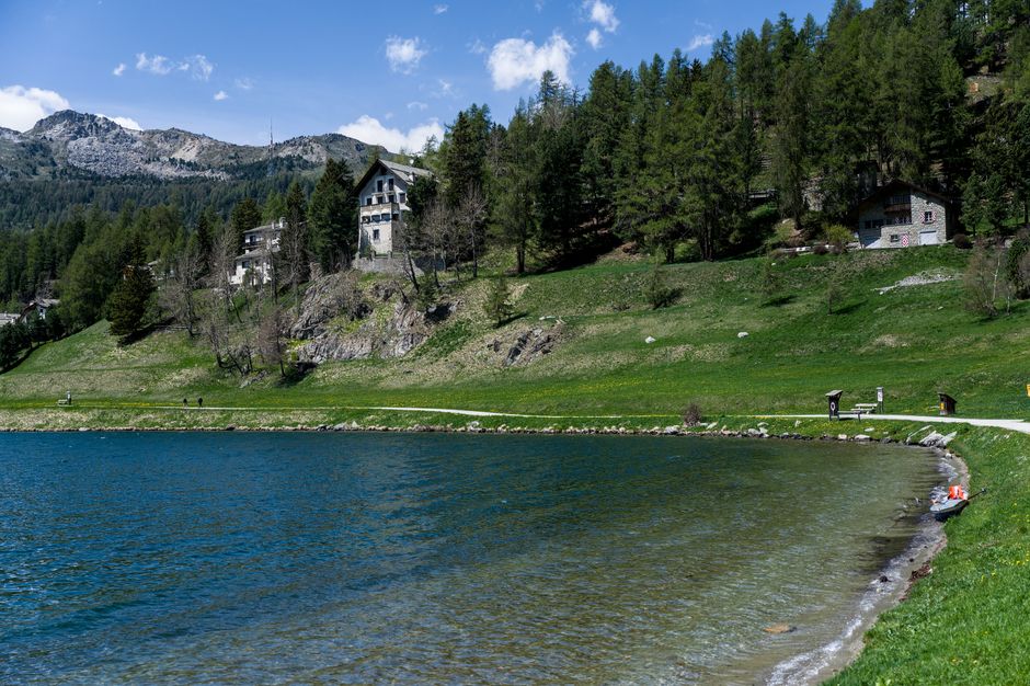 The image depicts a serene lakeside scene with a gently curving shoreline bordered by a narrow strip of dark, wet sand and small rocks. Clear, calm water with a slightly rippled surface extends across the lower part of the image, reflecting the bright blue sky overhead. Along the shoreline, there is a small, orange and white rubber boat resting on the grass near the water's edge. Behind the lake, a gently sloped grassy area features a few scattered small trees and shrubs, with a winding pathway that runs parallel to the water. Further back, several residential houses and cabins with muted grey or beige facades are nestled among dense, tall evergreen trees on a hillside. The backdrop reveals a rugged mountain range with jagged peaks under a partly cloudy sky, creating a tranquil, natural environment suitable for private outdoor recreation or landscape viewing. The scene is well-lit with natural daylight, emphasizing the lush greenery and the peaceful waters, highlighting the natural beauty of the area and its suitability for outdoor activities or scenic relaxation.